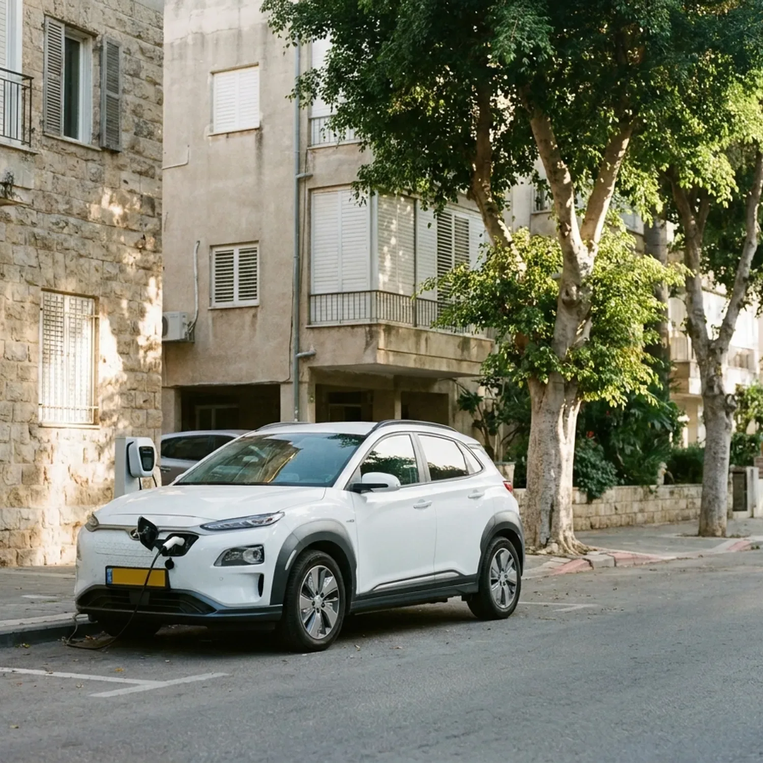 צילום פוטו-ריאליסטי של רכב פנאי חשמלי קומפקטי מודרני בצבע לבן חונה ברחוב עירוני ישראלי טיפוסי עם עצי פיקוס, מחובר לעמדת טעינה ביתית קטנה על קיר בניין, תאורת בוקר טבעית, ללא לוגו או טקסט על הרכב